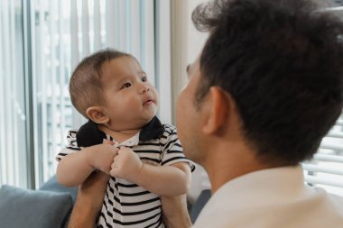 Bebeğini kucağında tutan bir babanın şefkatli anı. Bebek merakla ve güvenle yukarı bakıyor. Sıcak etkileşim aile sevgisini, yakınlaşmayı ve ebeveyn bakımını yansıtır..