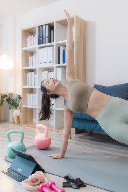 Kadın oturma odasında ileri yoga pozları veriyor, disiplin ve esneklik gösteriyor, fiziksel gücü, zihinsel odaklanmayı ve sağlığını günlük idmanlarla vurguluyor..