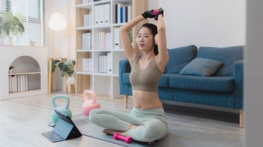 Genç bir kadın evde egzersiz yapıyor, yoga yapıyor ve halterleri ve dambılları ile egzersiz yapıyor. Egzersiz rutinini odaklanma ve kararlılıkla takip ediyor..