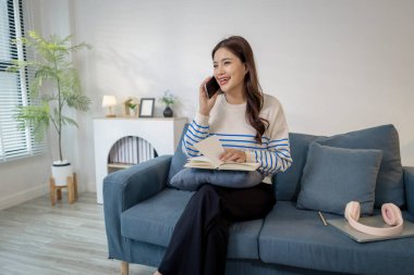 Genç bir kadın kanepede rahat bir şekilde oturur, elinde bir defter tutar ve akıllı telefonuyla gülerek konuşur. Rahatlamış görünüyor, notları okumak ve sohbet etmek arasında birden fazla iş yapıyor..