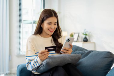 Genç bir kadın elinde akıllı telefon ve kredi kartıyla kanepede oturuyor. Çevrimiçi alışveriş yaparken gülümsüyor. Evde alışveriş yaparken rahat ve kendinden emin görünüyor..