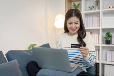 Genç bir kadın elinde bir kredi kartı tutarken bir dizüstü bilgisayar kullanarak kanepede rahatça oturuyor ve internet üzerinden ödeme yapıyormuş gibi görünüyor. Memnuniyetle gülümser, rahatlığı ve rahatlığı yansıtır..
