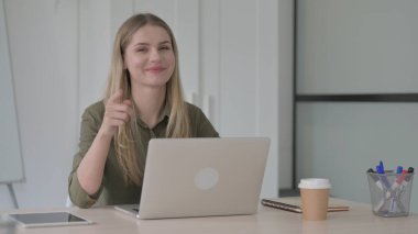 Ofiste dizüstü bilgisayar kullanırken kamerayı gösteren sarışın genç kadın.