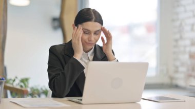 Baş ağrısı olan genç iş kadını dizüstü bilgisayarda çalışıyor.