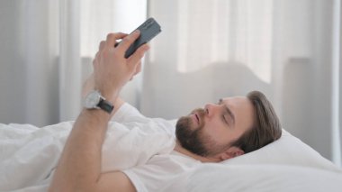Yatakta telefon kullanan Genç Yetişkin Adam Yan Görünümü