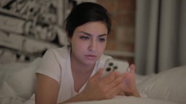 Genç kadın yatakta karnı üzerinde yatarken akıllı telefon kullanıyor.