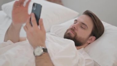 Genç Yetişkin Adam yatakta yatarken telefonu kullanıyor.