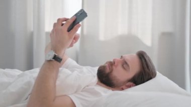 Yatakta telefon kullanan Genç Yetişkin Adam Yan Görünümü