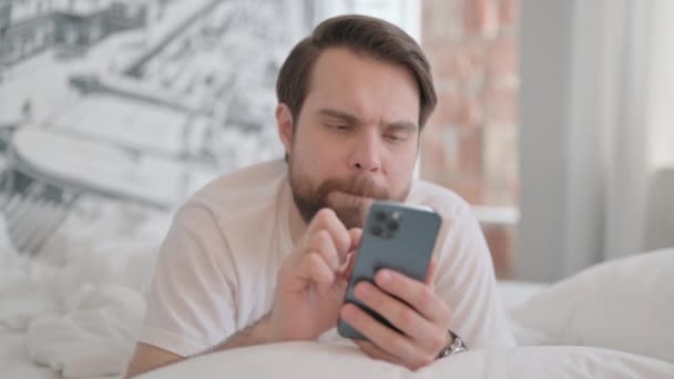 胃の上に横たわっている間 スマートフォンを使用して若い大人の男 — ストック動画