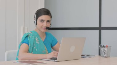 Kulaklıklı genç Hintli kadın Çağrı Merkezi 'nde kameraya doğru bakıyor.