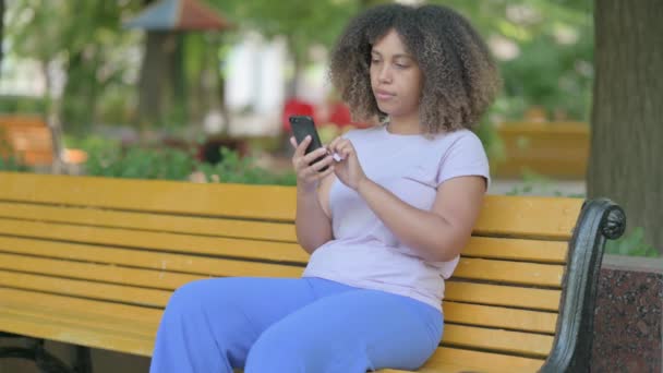 非洲女青年坐在户外长椅上 一边用智能手机庆祝成功 — 图库视频影像