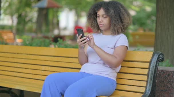 非洲年轻女子坐在室外的长椅上 在智能手机上丢失而震惊 — 图库视频影像