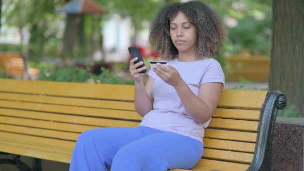 年轻的非洲妇女坐在户外长椅上成功地通过电话购物而感到兴奋 — 图库视频影像