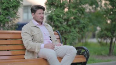 Orta yaşlı adam baş ağrısıyla dışarıda oturuyor.