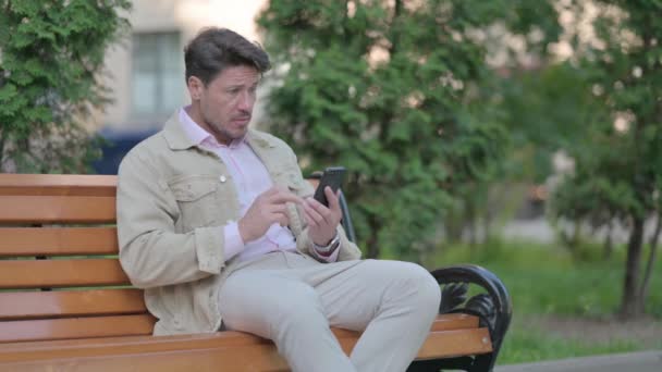 Medelålders Man Chockad Förlust Smartphone Medan Sitter Utomhus Bänk — Stockvideo