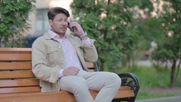 ベンチに座っている間に電話で主張する中年男性の動揺 — ストック動画