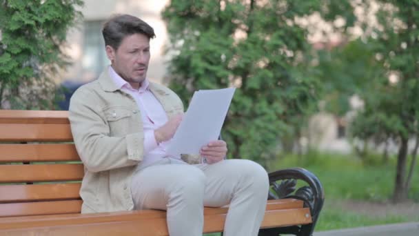 ベンチで屋外に座っている間に中年男性読書契約の動揺 — ストック動画