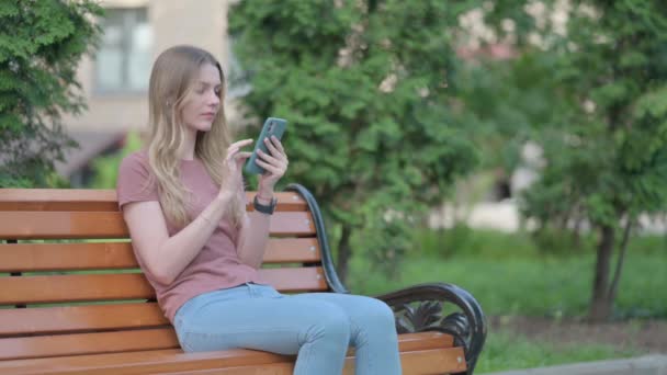 年轻女子坐在户外的长椅上 在智能手机上丢失而震惊 — 图库视频影像
