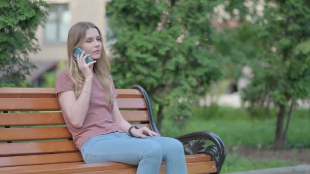 坐在户外长椅上打电话聊天的年轻女人 — 图库视频影像