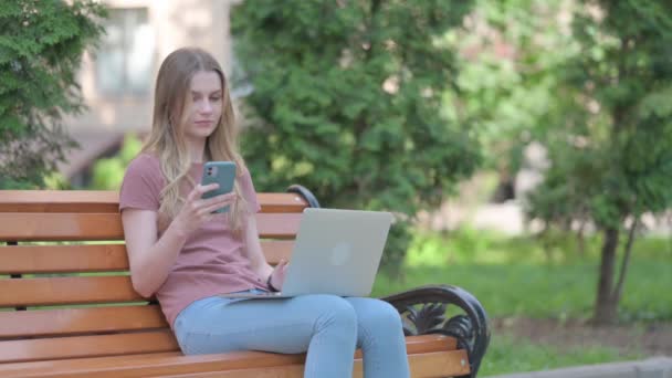 Młoda Kobieta Korzystanie Laptopie Smartfonie Podczas Siedzenia Zewnątrz Ławce — Wideo stockowe