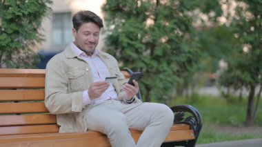 Orta Yaşlı Adam Bir Benç 'te Açık Otururken Telefonla Başarılı Bir İnternet Alışverişi İçin Heyecanlı