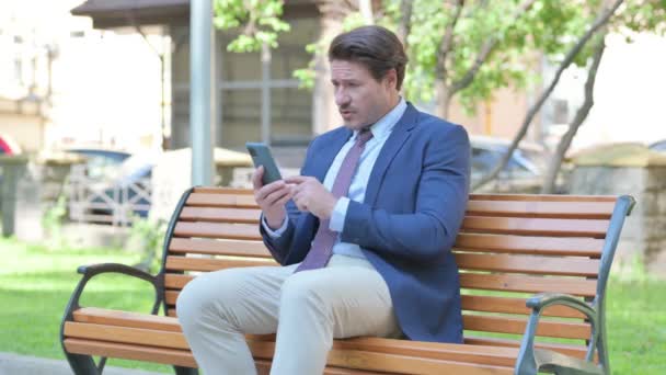 中年のビジネスマンは ベンチに屋外に座っている間 スマートフォンで損失にショックを受けました — ストック動画