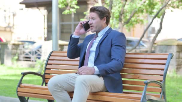 Empresário Meia Idade Perturbado Discutindo Telefone Enquanto Sentado Livre Banco — Vídeo de Stock