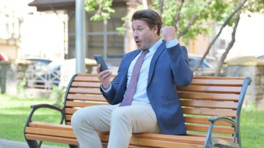 Smartphone 'da Başarı Kutlaması Yapan Orta Yaşlı İşadamı Bir Bankta Açık Otururken