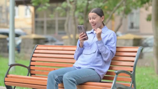 Latynoska Kobieta Świętuje Sukces Smartfonie Siedząc Zewnątrz Ławce — Wideo stockowe