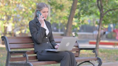 İş kadını dışarıda dizüstü bilgisayar kullanırken telefonda konuşuyor