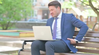 Sırt ağrısı olan bir iş adamı Bench 'te otururken dizüstü bilgisayar kullanıyor.