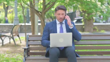 Orta yaşlı iş adamı telefonda konuşuyor ve dışarıda dizüstü bilgisayar kullanıyor.