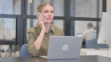 Genç iş kadını dizüstü bilgisayarla çalışırken telefonla konuşuyor.