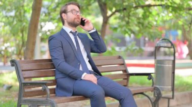 Kızgın sakallı iş adamı dışarıda otururken telefonda konuşuyor.