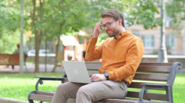 Migreni olan genç adam dışarıda dizüstü bilgisayar kullanıyor.