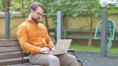 Orta yaşlı adam dışarıda otururken dizüstü bilgisayar kullanıyor.