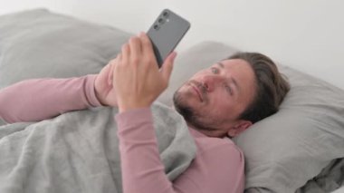Orta yaşlı adam yatakta yatarken akıllı telefon kullanıyor.