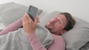 Orta yaşlı adam yatakta yatarken telefonda görüntülü konuşma yapıyor.