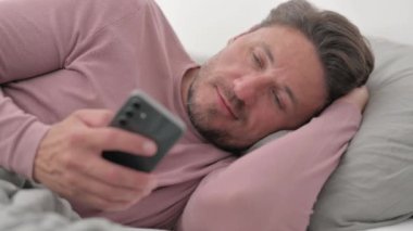 Orta yaşlı adam yatakta yan yatarken telefonu kullanıyor.