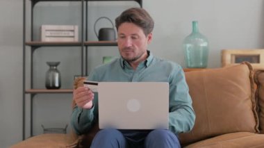 Orta yaşlı adam internette kucağında dizüstü bilgisayarla alışveriş yapıyor.