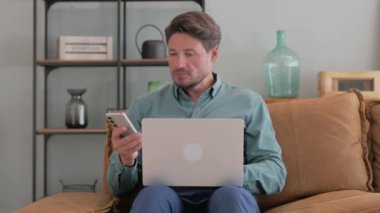 Orta yaşlı adam dizüstü bilgisayarlı telefonu kullanıyor.