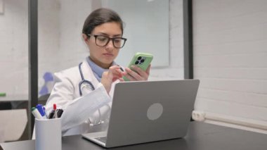 Klinikte telefon ve dizüstü bilgisayar kullanan Hintli genç doktor.