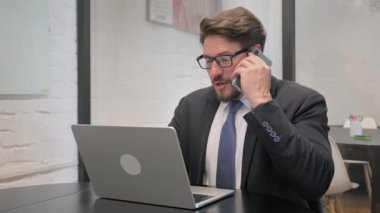 Orta yaşlı iş adamı telefonda konuşuyor ve dizüstü bilgisayarında daktilo kullanıyor.