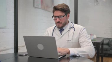 Başağrısı olan orta yaşlı bir doktor dizüstü bilgisayarda çalışıyor.