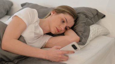 Yatakta yan yatarken telefonu kullanan genç kadın.