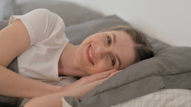 Yatakta yatarken kameraya bakan gülümseyen genç kadın.