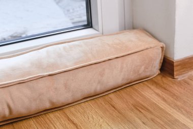 Draft excluder lying in front of door to keep out cold air and save energy for heating in room clipart
