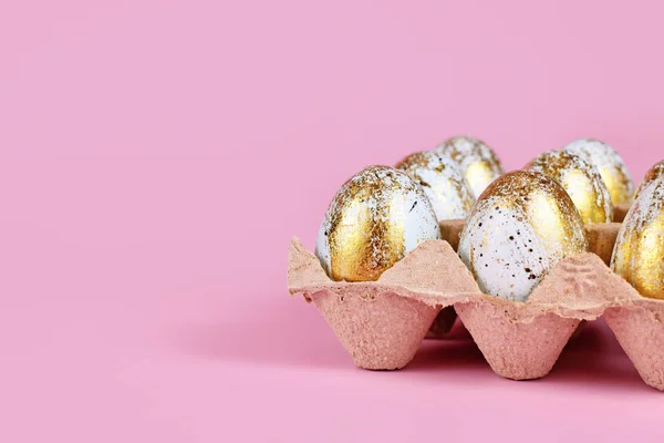 Painted blue Easter eggs with golden spots in egg carton on pink background with copy space