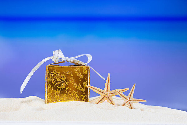 Golden packed gift box with yellow stars at tropical ocean beach. Christmas, New Year, birthday vacation in hot countries background concept. Copy space