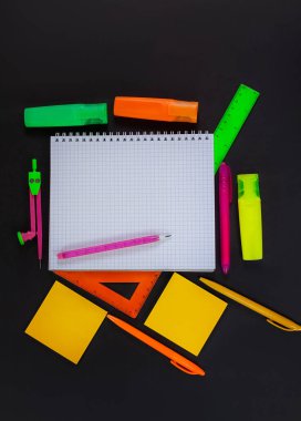 Okula dönelim. Siyah masanın üzerinde boş boşluğu olan kalemle işaretlenmiş bir defter var. Çevrede yeşil, sarı, pembe, turuncu kırtasiye malzemeleri var. Cetvel, keçeli kalem, kalem, yapıştırıcı. Üst görünüm